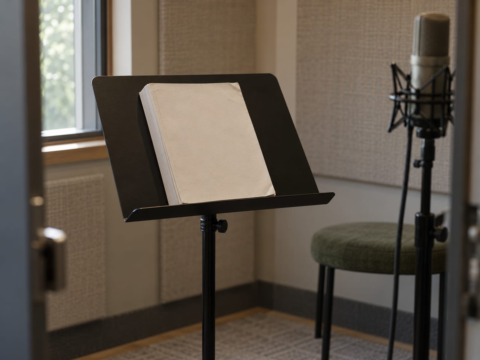 A single microphone in a small recording booth beside one music stand and a thick closed manuscript with a plain cover