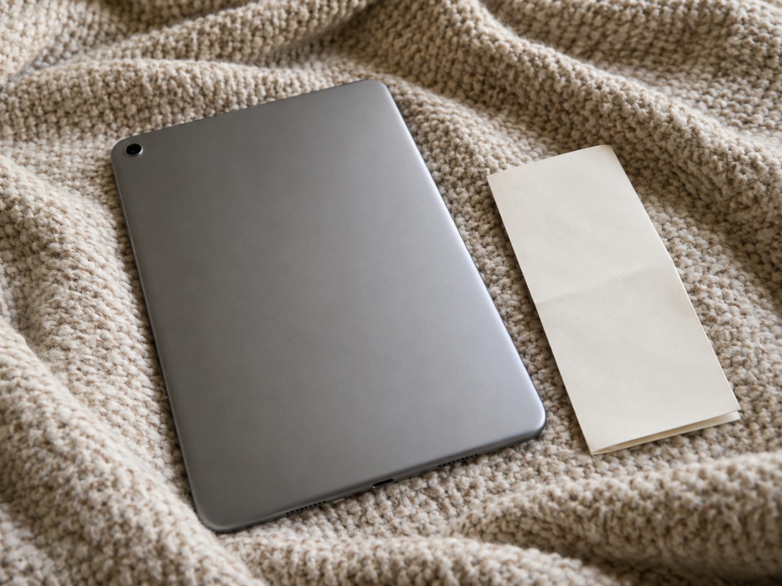 An unbranded tablet lies face down on a bed cover beside a folded blank paper slip shaped like a receipt.